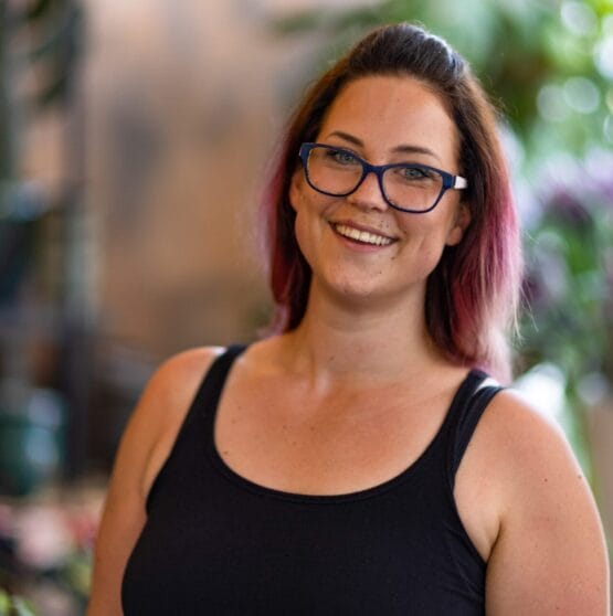 Eine Frau mit Brille lächelt vor einer Topfpflanze. {{brizy_dc_image_alt imageSrc=