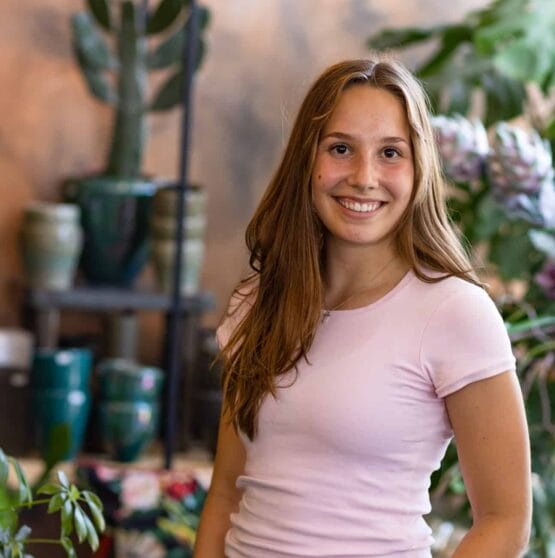 Eine junge Frau in einem rosa Hemd steht vor Topfpflanzen.