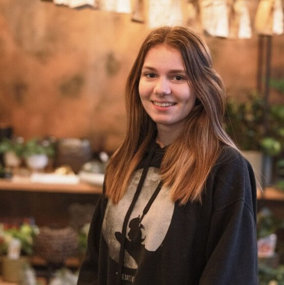 Eine junge Frau in einem schwarzen Kapuzenpullover steht vor Pflanzen. {{brizy_dc_image_alt imageSrc=